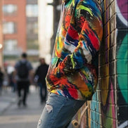 Person wearing a colorful hoodie leaning against a graffiti-covered wall on a street.