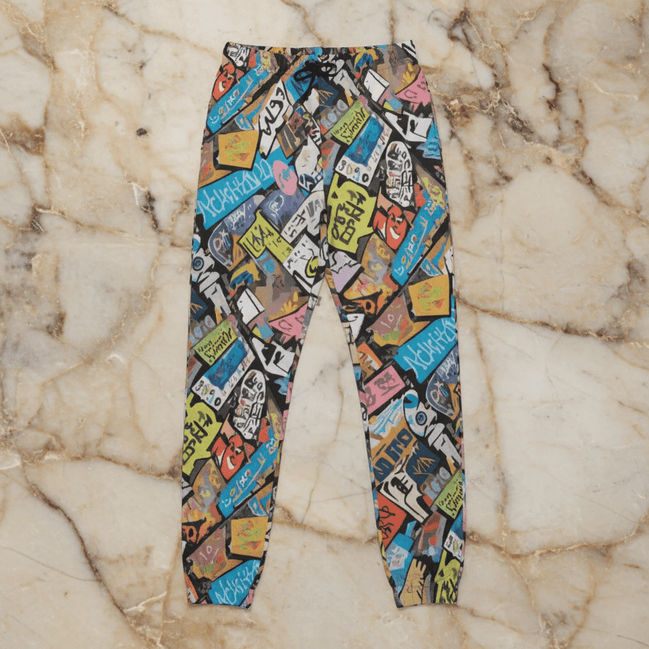 Tag Stack Riot lounge pants by Dyles Mavis — heavyweight cotton sweatpants with all-over print of a dense street-sticker and graffiti tag collage in blue, pink, yellow and black, flat lay on marble