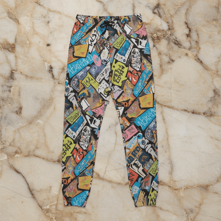 Tag Stack Riot lounge pants by Dyles Mavis — heavyweight cotton sweatpants with all-over print of a dense street-sticker and graffiti tag collage in blue, pink, yellow and black, flat lay on marble