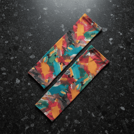 Boundless Bleeds UV arm sleeves by Dyles Mavis — cooling lycra arm sleeves with all-over print of abstract expressionist paint splatter in teal, red, orange and black, flat lay on dark stone