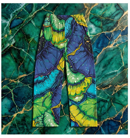 Butterfly Wing pajama pants by Dyles Mavis — brushed polyester lounge pants with all-over print of emerald and sapphire organic butterfly wing scale patterns and gold vein detailing, flat lay on green marble