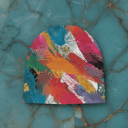 Painted Camo Knit Beanie in soft stretch knit with multicolor brush-dab pattern in crimson, teal, orange, magenta, and green with spiky splatter accents, flat lay on teal marble
